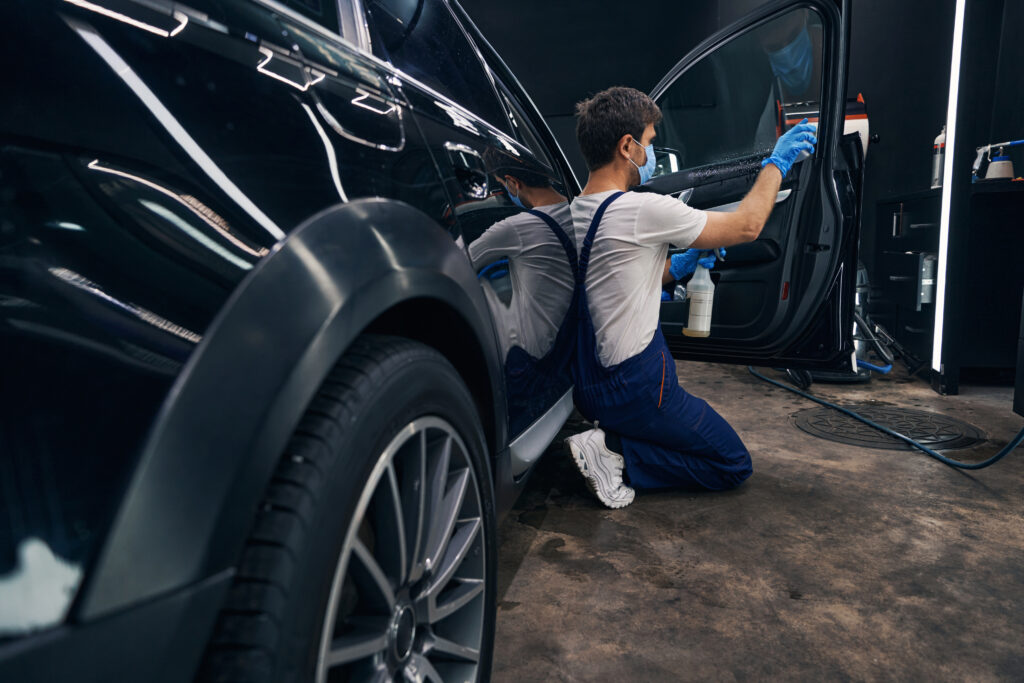 Car mechanic standing on knees and doing car valeting by washing its window with cleaning spray