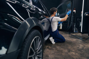 Car mechanic standing on knees and doing car valeting by washing its window with cleaning spray